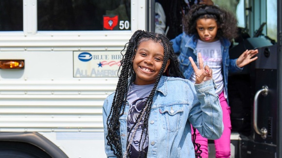A child holds up a peace sign as she walks off a school bus